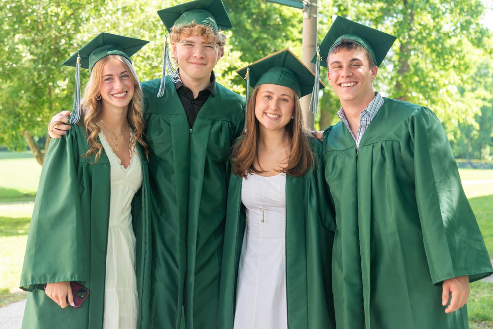 Four graduates (two female two male) smiling stand close together wearing green caps and gowns with trees behind them